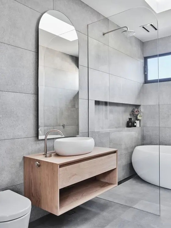a contemporary grey bathroom with large scale tiles, an oval tub and a shower space, a stained vanity, an arched mirror