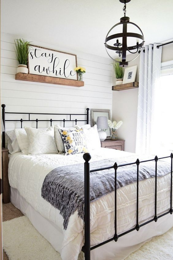 a cute modern farmhouse bedroom with white wooden plank walls, a forged bed, a sphere chandelier, shelves and greenery