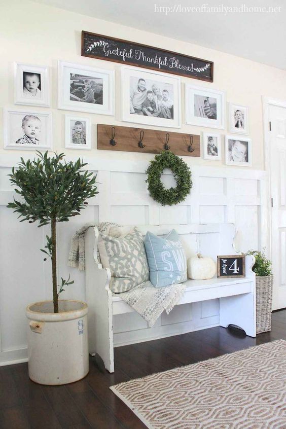a farmhouse entryway with a white bench with pillows, a pumpkin, a gallery wall, a greenery wreath and a potted tree
