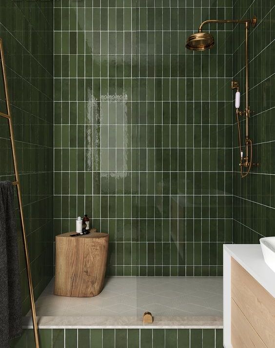 a green bathroom clad with long and narrow tiles, with gold and brass touches and a tree stump stool in the shower