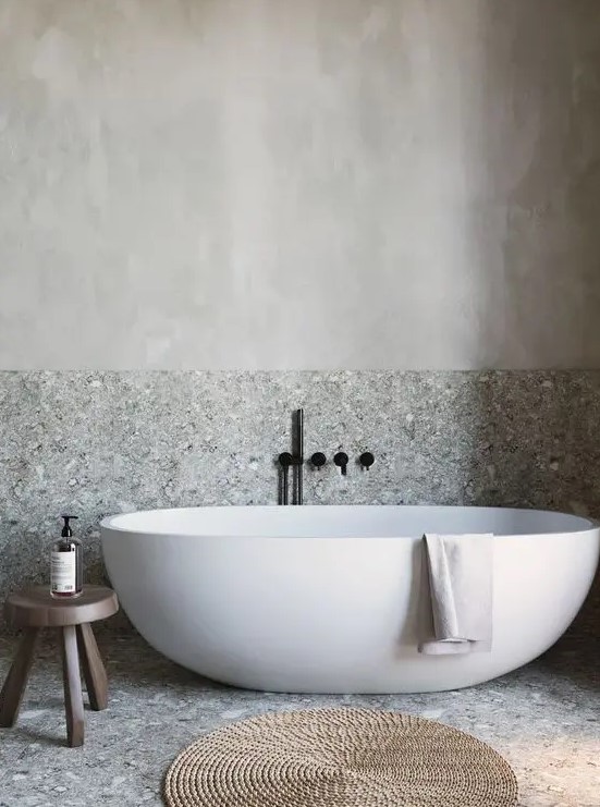 a grey bathroom done with stone and plaster, with a free standing tub, a wooden stool and a jute rug