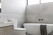 a grey minimalist bathroom clad with large scale tiles, an oval tub, a long and narrow window and a floating vanity