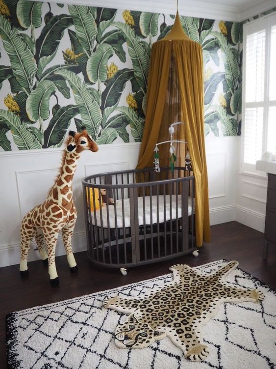 a jungle nursery with a tropical leaf wall, a dark crib, a mustard canopy, a fun rug and a toy giraffe