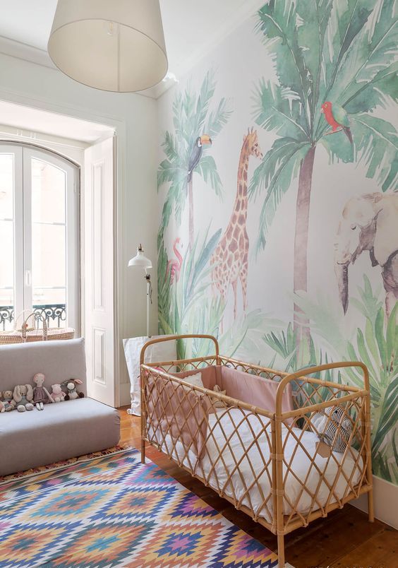 a jungle themed nursery with a statement wall, fun toys, a printed rug and a rattan crib with pastel bedding