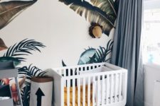 a jungle themed nursery with tropical leaf prints on the wall, a polka dot rug, blue curtains, a unique chandelier and colorful bedding