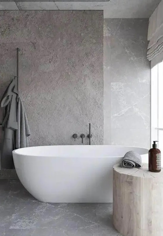 a minimalist and refined grey bathroom fully clad with stone, with a large tree stump side table and an oval tub is chic