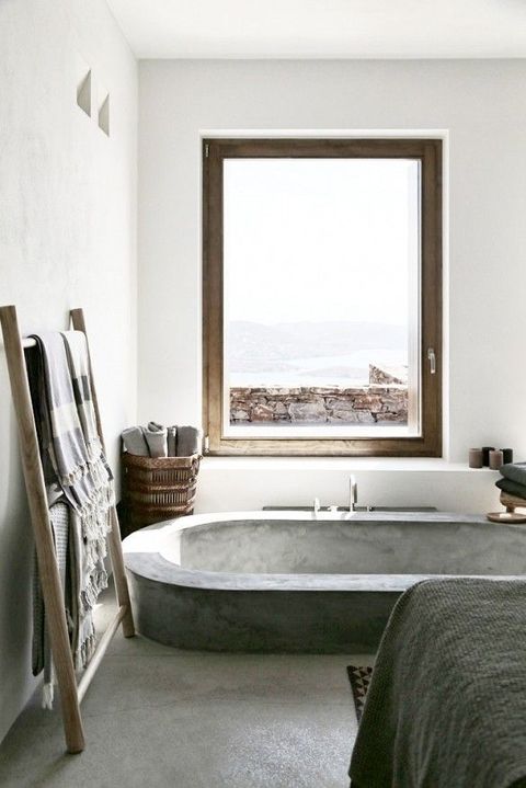 a minimalist industrial bathroom with a concrete floor and bathtub, touches of wood and wicker, textiles and a large window for views