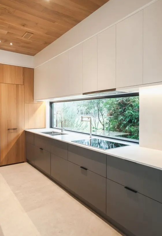 a minimalist kitchen with lower grey and upper white cabinets, a window backsplash, a white countertop and stained wood for a warmer feel
