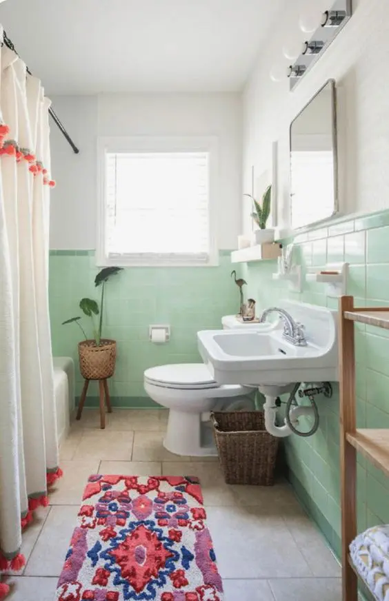 a mint green bathroom with a wlal mounted sink, a toilet and a bathtub, a bold rug and a pompom curtain