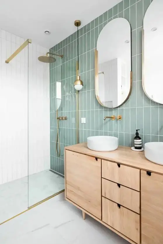 a modern bathroom with a sage green tile wall, a stained vanity, oval mirrors and round sinks, a shower space enclosed in glass
