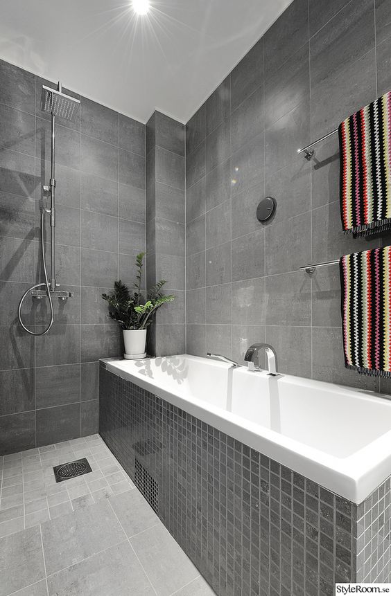 a modern grey bathroom clad with various types of tiles, a bathtub clad with tiles, colorful towels and a potted plant