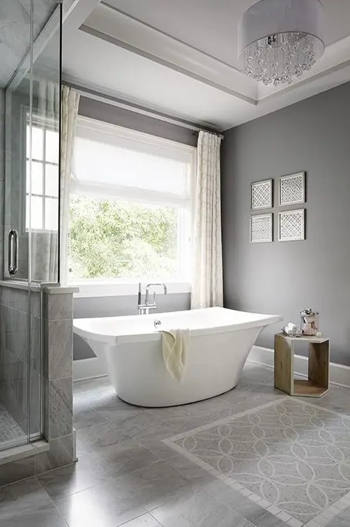 a modern grey bathroom done with tiles on the floor, a catchy free standing tub, a wooden table and a crystal chandelier