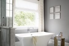 a modern grey bathroom done with tiles on the floor, a catchy free-standing tub, a wooden table and a crystal chandelier