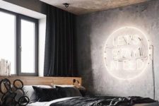 a modern industrial bedroom with a concrete ceiling and walls, a neon light, a platformbed and dark textiles