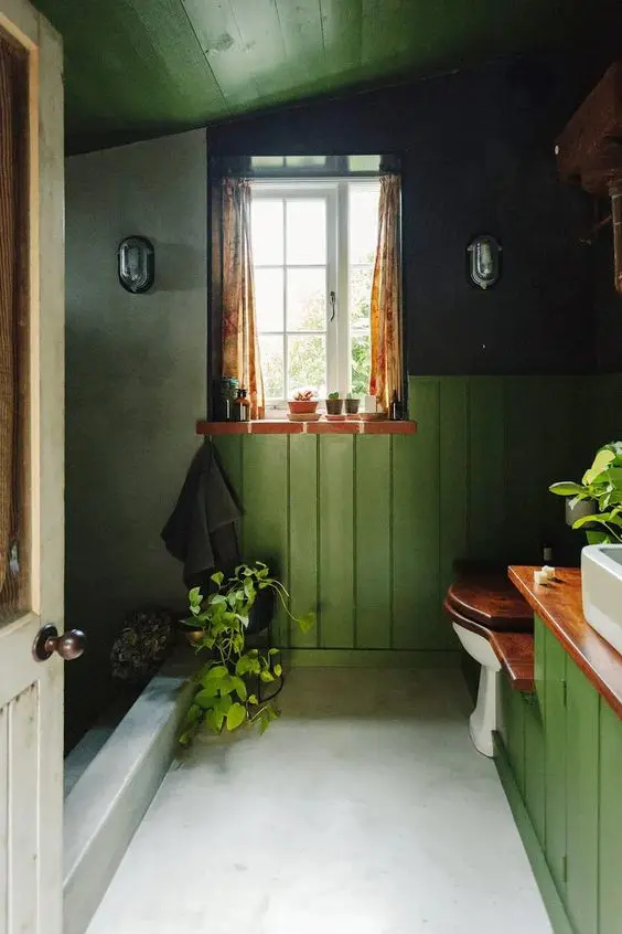 a pretty vintage bathroom with green paneled walls and a ceiling, a shower space, a window and a green vanity plus some plants