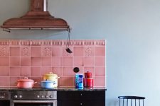 a quirky kitchen with black cabinets, a bright pink tile backsplash, a copper hood and a pink pendant lamp