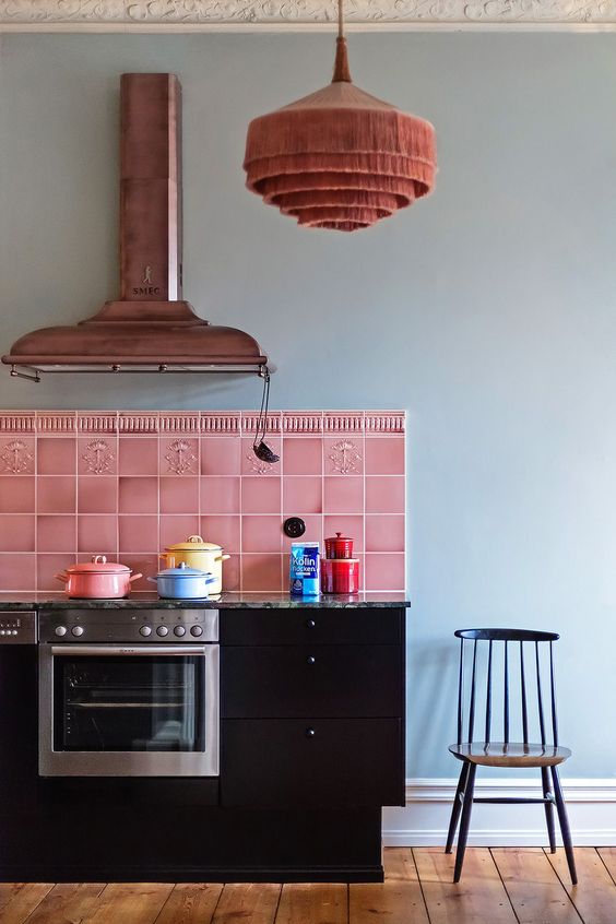 a quirky kitchen with black cabinets, a bright pink tile backsplash, a copper hood and a pink pendant lamp
