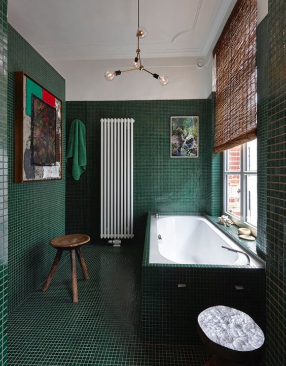 a refined dark green bathroom all clad with tiles including the tub, a modern chandelier, a wooden stool and some pretty artworks