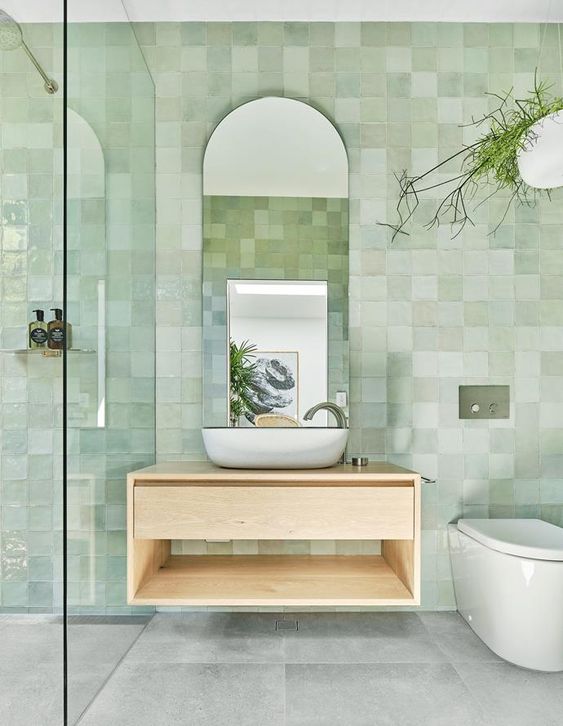 a serene bathroom clad with light green glazed tiles, a shower space, a stained vanity, an arched mirror and potted plants