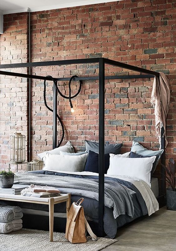 a simple industrial bedroom with red brick walls, a black metal frame bed, wooden and wicker furniture and cozy textiles