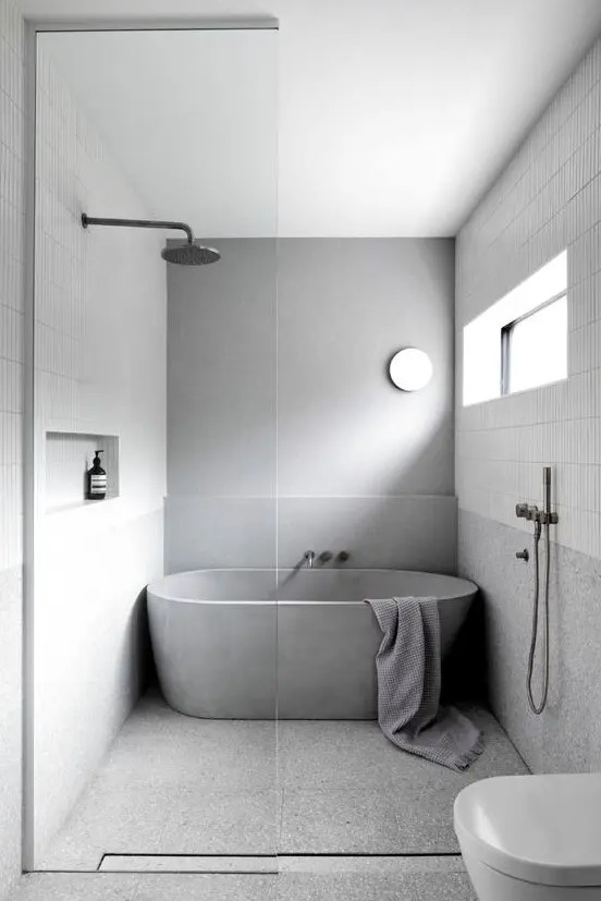 a simple minimalist bathroom clad with skinny white tiles and grey stone-like ones, a concrete tub, white appliances