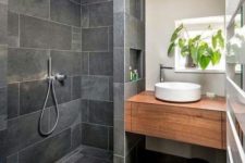 a small grey bathroom clad with tiles, with a built-in wooden vanity, white appliances and potted greenery