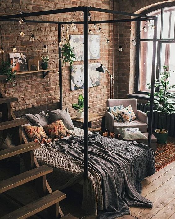 a small yet chic industrial bedroom with red brick walls, a canopy bed, potted greenery and lights plus catchy textiles