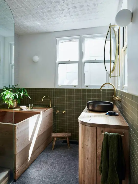 a spa style bathroom clad with green square tiles, with a wooden ofuro tub and a wooden vanity, a black sink and gold fixtures