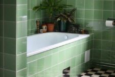 a stylish bathroom with a black and white floor and green tiles on the walls and cladding the tub, potted greenery is a cool space