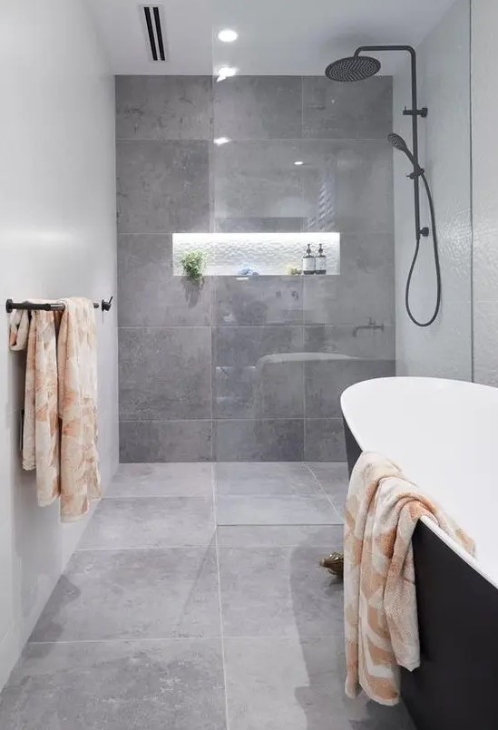 a stylish contemporary grey bathroom clad with large scale tiles, with a lit up niche, a black tub and dark fixtures