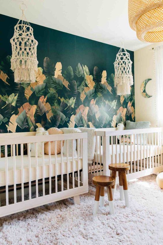 a tropical nursery with a trendy dark tropical leaf wall, boho chandeliers, painted stools and fun pillows