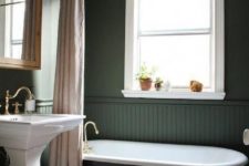 a vintage dark green bathroom clad with beadboard, a black tub, a free-standing sink, pastel textiles and touches of brass