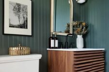 a vintage powder room with dark green shiplap, a stained vanity, a toilet, an arched mirror, some gold and brass touches