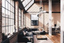 an airy industrial living room with windows and skylights, brick walls, a wooden ceiling, comfy furniture and a catchy chandelier