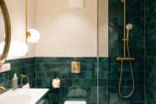 an elegant bathroom clad with dark green tiles and a dark green vanity, a mirror, a toilet and a shower space, some gold fixtures