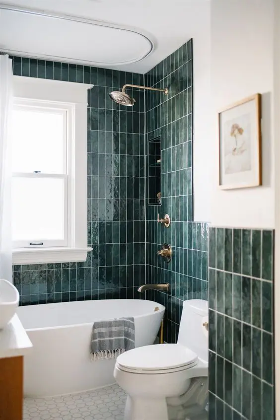 an elegant bathroom with dark green skinny tiles and white hex tiles, an oval tub and a toilet, a stained wood vanity and neutral fixtures