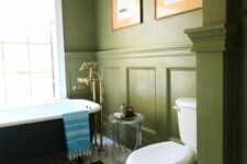 an elegant vintage bathroom with grass green paneled walls, a black bathtub, a white toilet, geo tiles on the floor and a gold chandelier