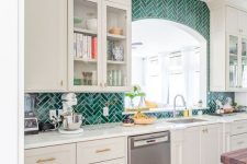 an elegant white kitchen with farmhouse cabinets, a bold emerald herringbone tile backsplash and white countertops
