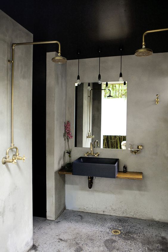 an industrial bathroom with concrete walls and a floor, with hanging bulbs, brass and gold fixtures and a floating vanity