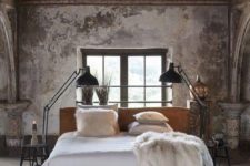 an industrial bedroom with shabby chic walls, a wooden ceiling, a concrete floor and upholstered bed, metal floor lamps and a vintage suitcase
