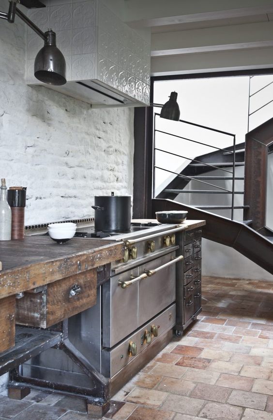 an industrial kitchen with rough wooden cabinets, metal appliances, a tile clad hood and vintage lamps