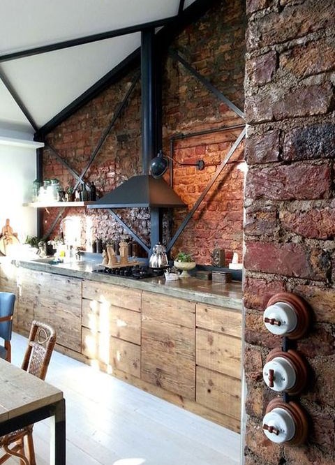 an industrial kitchen with rough wooden furniture with concrete countertops, brick walls, a black hood, mismatching furniture