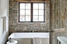 a chalet bathroom with white walls and a stone one, wooden beams, a modern tub and stool and a reclaimed wooden door