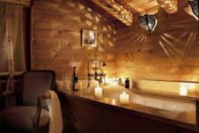 a chic chalet bathroom clad with light-colored wood completely, with a wood clad tub, refined furniture and lights and candles