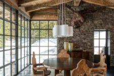 a chic chalet dining room with a stone wall, a faux deer head, a vintage table and rustic chairs plus a glazed wall