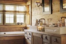 a cozy chalet bathroom clad with light-colored wood, with a large vanity and a wood clad bathtub, windows and mirrors