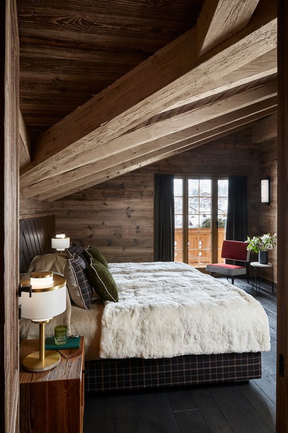 a cozy chalet bedroom clad with wood, with upolstered furniture, chic lamps and an entrance to the balcony