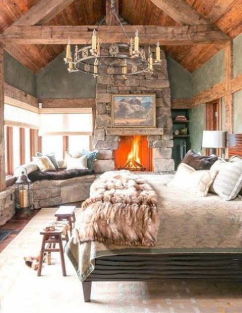 a cozy chalet bedroom with a stone clad fireplace, a vintage bed, a vintage chandelier, a stone bench and wooden stools