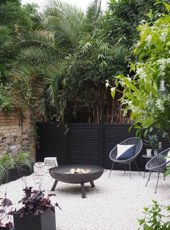 a laconic modern backyard with white gravel, a black fire pit, woven chairs, potted greenery and other plants