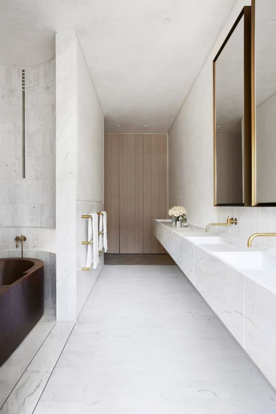 a large minimalist bathroom done with white stone, a large floating vanity, a mirror in a gilded frame and a dark tub
